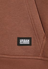 Pruun kapuutsiga, millel on tekstureeritud kangas, esiküljel on must silt, millel on julged valged tähed "URBAN CLASSICS".