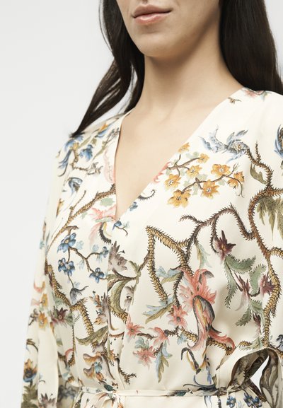 Γυναίκα που φοράει κρεμ μπλούζα με λουλουδάτα και κλαδιά σε απαλούς μπλε, πορτοκαλί, ροζ και πράσινους τόνους.