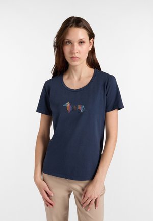 Młoda kobieta z długimi brązowymi włosami ubrana w granatowy t-shirt z kolorowym, paskowanym wzorem jamnika i beżowe spodnie na białym tle.