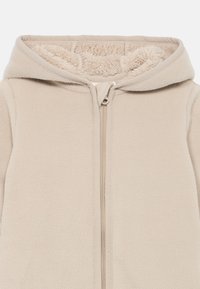 Beige fleece jack met een capuchon, voorzien van een volledige rits en een zachte, tekstuurlijke voering aan de binnenkant. Naaddetails langs de rits.