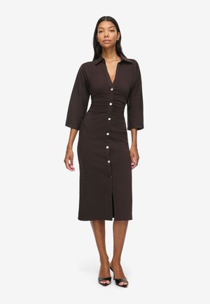 Femme portant une robe midi marron foncé à boutons sur le devant, manches trois quarts et talons noirs ouverts, debout devant un fond blanc uni.