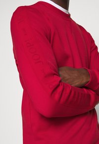 Sudadera roja de algodón, con cuello redondo y detalle de logotipo elevado a lo largo de las mangas. Textura suave con un diseño ajustado.