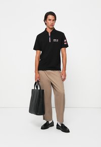 Sort polo t-shirt med farvet krave og USA-logoer, parret med beige bukser og en sort læder tote bag. Modellen bærer sorte sko.