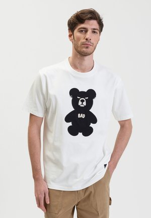 Witte katoenen t-shirt met een zwarte beer graphic en het woord "BAD" op de borst. Korte mouwen en ronde hals. Eenvoudig ontwerp.