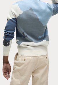 Sweter z przędzy z górskim wzorem w odcieniach niebieskiego, białego i zielonego. Posiada ściągacze na mankietach i u dołu, zestawiony z jasnobezowymi spodniami z korduroyu.