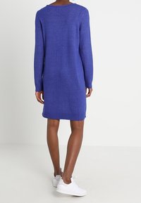 Persona che indossa un vestito a maglia viola con maniche lunghe e lunghezza al ginocchio, abbinato a scarpe da ginnastica bianche, posizionata di fronte a uno sfondo bianco semplice.