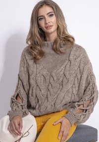 Brązowy sweter robiony na drutach z splotami warkoczowymi i szerokimi rękawami. Posiada wycięcia na łokciach. Połączony z żółtymi spodniami z korduroy.