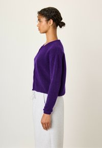 Vijolični skrajšani cardigan iz puhastega materiala z dolgimi rokavi, okroglo ovratnico in zapenjanjem s gumbi spredaj.