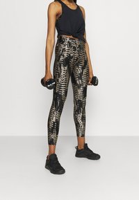 Černý top na ramínka uvázaný v pase, doplněný o vzorované, přiléhavé legíny ve zlaté a černé barvě. Modelka drží činky a má na sobě černé tenisky.