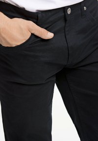 Zwarte broek heeft een slim design met een voorzak. De stof lijkt glad en er is een metalen knoop zichtbaar bij de taille.