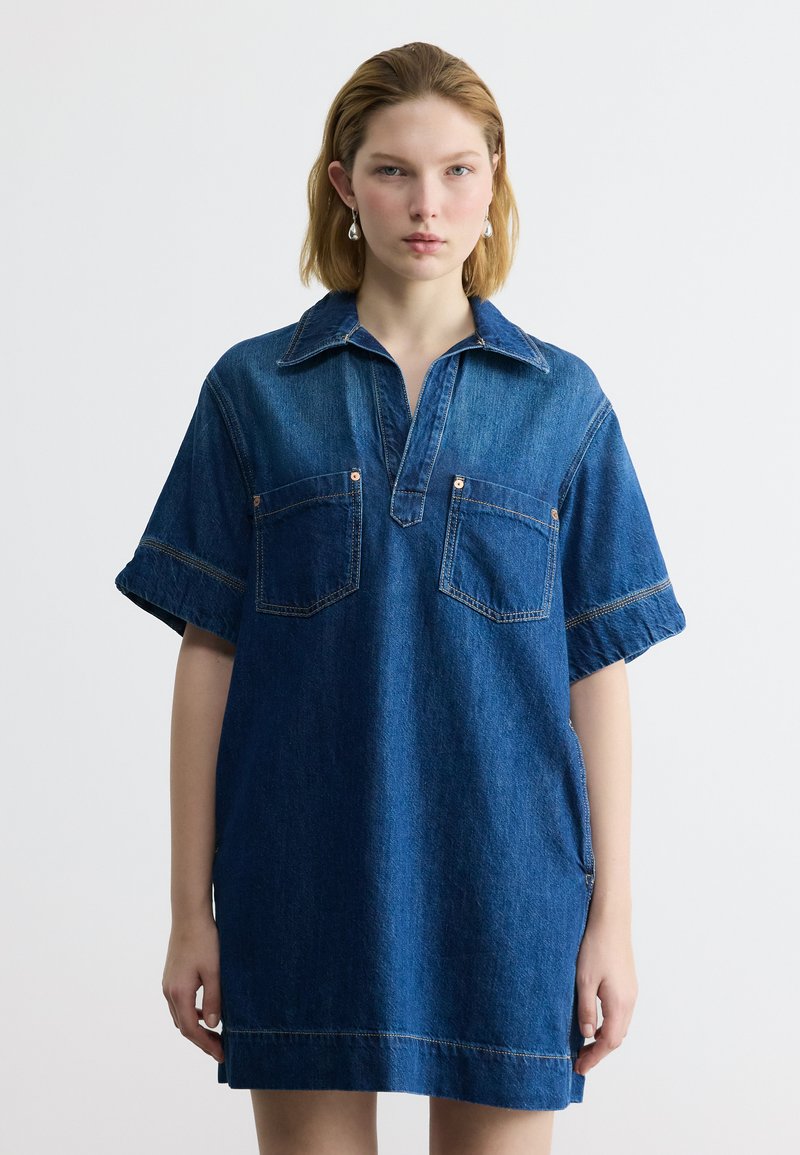 Jeune femme portant une robe en jean ample à manches courtes avec col et deux poches frontales, debout devant un fond clair uni.