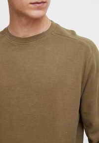 Chemise longue à manches longues de couleur vert olive, texturée avec un motif côtelé, col rond et coutures subtilement brodées.