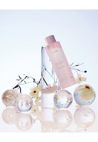 Rosa flaska med Huxley hudvård i guldtext, placerad mot en klar glasvas och sfäriska dekorationer, med fina blommor och reflektioner.