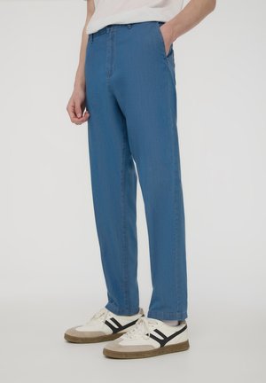 Persona che indossa pantaloni casual blu con tasche laterali e sneakers bianche con strisce nere, in piedi davanti a uno sfondo semplice.