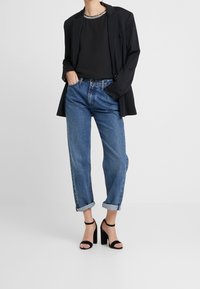Blazer negro sobre una blusa negra, combinado con jeans de mezclilla claros enrollados y sandalias de tacón alto negras. Diseño elegante con un toque casual.