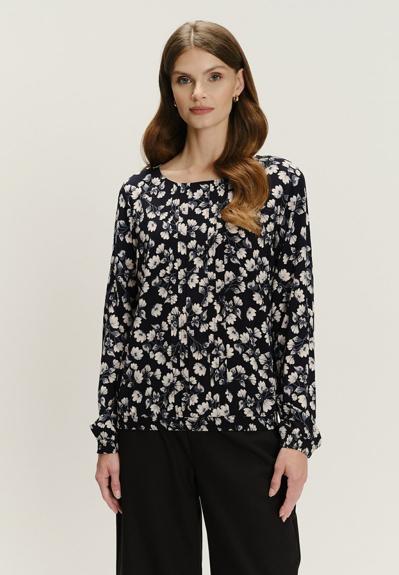 Frau mit langen, gewellten Haaren, die eine marineblaue, blumige Bluse mit langen Ärmeln und schwarze Hosen trägt, steht vor einem schlichten, hellen Hintergrund.