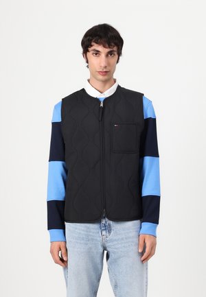 Young man wearing black quilted vest over blue and navy striped long sleeve shirt and light blue jeans, standing against a plain white background.
