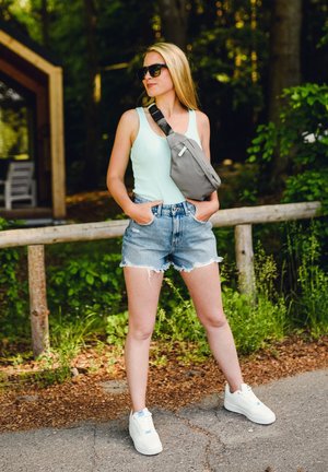 Lässiges Outfit mit einem hellgrünen Tanktop, zerrissenen Jeansshorts und weißen Sneakers. Eine graue Umhängetasche hängt über der Schulter.