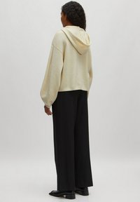 Beige, kürzer geschnittenes Hoodie mit Kapuze, strukturierter Stoff und weiten Ärmeln, kombiniert mit schwarzen, weit geschnittenen Hosen und schwarzen Schuhen. Rückansicht.