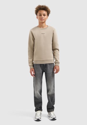 Teenager mit lockigem Haar, trägt einen beigen Sweatshirt, ausgebleichte schwarze Jeans und weiße Sneakers, steht mit neutralem Ausdruck vor einem hellen Hintergrund.