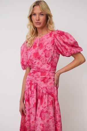 Moteris vilki rožinę gėlėtą suknelę su pūstais rankovėmis, laiko tekstūruotą rožinės spalvos rankinę, stovėdama prieš vienodą foną.