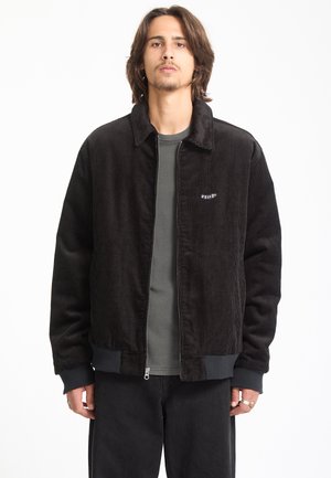 Jeune homme aux cheveux mi-longs porte une veste zippée en velours côtelé noire par-dessus une chemise gris foncé sur un fond blanc uni.