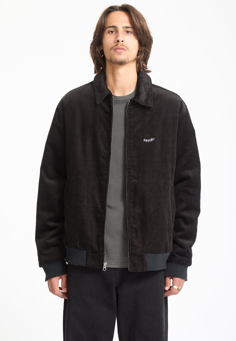 Jeune homme aux cheveux mi-longs porte une veste zippée en velours côtelé noire par-dessus une chemise gris foncé sur un fond blanc uni.