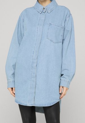 Person iført en lys blå oversized denimskjorte med en brystlomme foran og sorte bukser mod en ensfarvet baggrund.