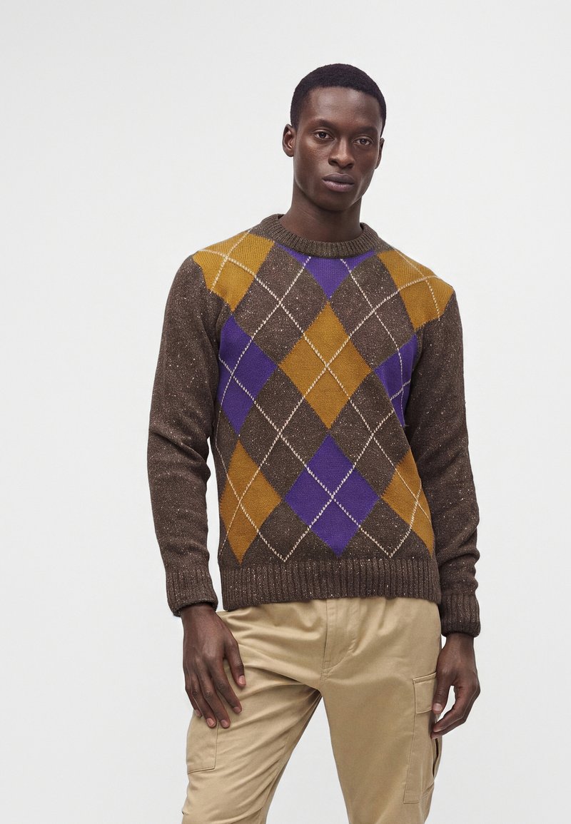 Homme portant un pull marron avec un motif losange moutarde et violet et un pantalon beige, se tenant devant un fond uni.