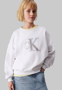 Biela oversized mikina so striebornou výšivkou loga "CK", okrúhlym výstrihom a rebrovanými manžetami, nosená cez žlté tričko a džínsovú sukňu.