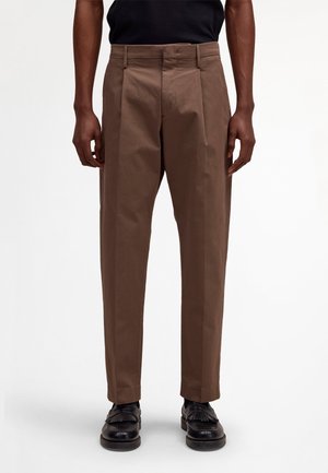 Mężczyzna noszący brązowe spodnie plisowane i czarne skórzane loafersy, stojący na tle jednolitego białego tła.