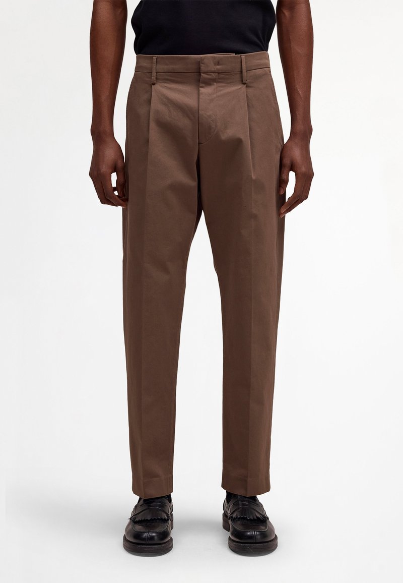 Homme portant un pantalon plissé marron et des mocassins en cuir noir, debout devant un fond blanc uni.