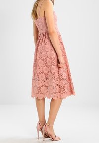 Robe en dentelle rose pâle, sans manches, avec un corsage ajusté et une jupe évasée, ornée de motifs floraux et d'un ourlet en forme de vagues. Assortie à des talons roses.