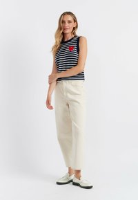 Top senza maniche a righe navy e bianche con un dettaglio a forma di cuore rosso, abbinato a pantaloni larghi color crema e scarpe di colore chiaro.