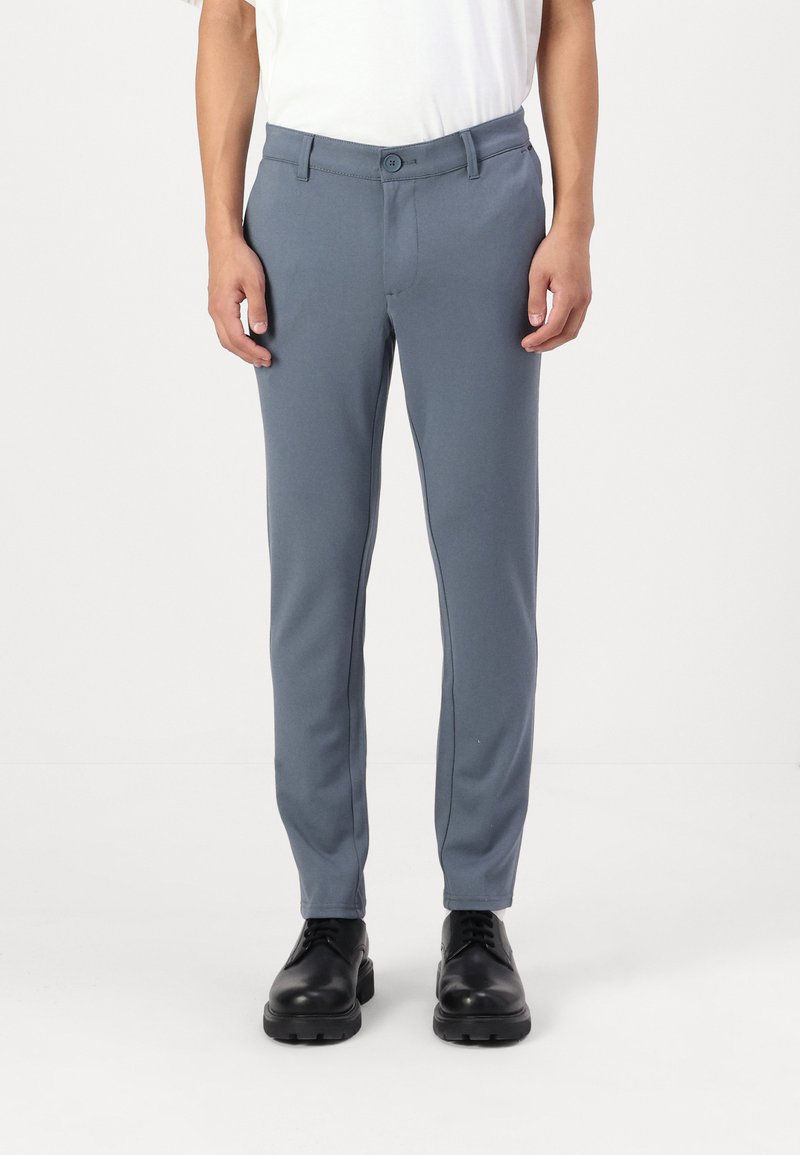 Pantalones formales de color azul claro hechos de un material suave y elástico. Presentan un frente plano, pasadores para cinturón y un corte slim. Se llevan con zapatos negros.