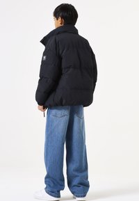 Chaqueta de plumas negra con un cuello alto, diseño acolchado y un parche del logo en la manga. Combinada con jeans azules holgados y zapatillas blancas.