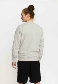Man met een bun-haarstijl, gekleed in een lichtgrijze sweatshirt met lange mouwen en zwarte shorts, staand met zijn gezicht weg van de witte achtergrond.