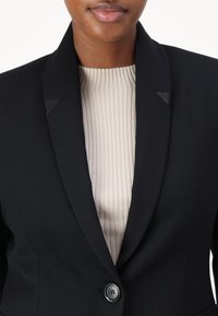 Crni krojeni sako s oštrim reverima, zatvaranjem na jedan gumb i suptilnim teksturiranim detaljima, nosi se preko rebraste bež majice.