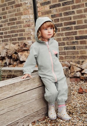 Young child in a light gray hooded jumpsuit with a pink zipper and floral rain boots, leaning on wooden planter outdoors.