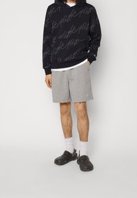 Schwarzer Hoodie mit einem wiederholten Schriftmuster, graue Shorts, weißes Unterhemd und schwarze Clogs mit Lüftungslöchern und Fersenriemen.