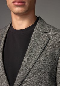Grå hørternet blazer over en sort crew-neck t-shirt. Blazeren har klaplugter og en blød tekstur, med synlige syninger.