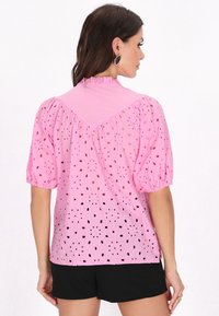 Rosa Kurzarmbluse mit Puffärmeln, die Ausschnittmuster und einen hohen Kragen aufweist. Der Stoff wirkt leicht und strukturiert. Schwarze Shorts sind sichtbar.