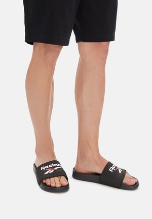 Osoba nosząca czarne klapki Reebok i czarne szorty, stojąca na jednolitym białym tle.