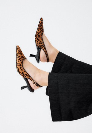 Piernas en pantalones negros que llevan zapatos slingback de tacón kitten con estampado de leopardo y puntas afiladas, levantadas contra un fondo blanco.