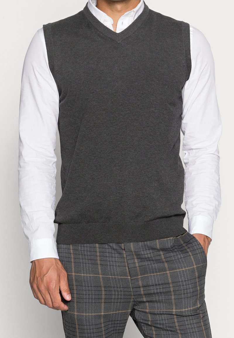 Homme portant un pull sans manches gris foncé à col en V sur une chemise blanche avec un pantalon à carreaux gris, main dans la poche.