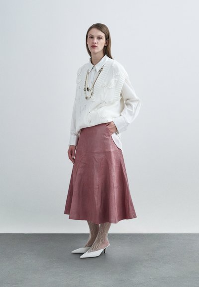 Pull en maille blanche avec motif floral sur une chemise blanche, associé à une jupe midi en cuir rose. Le mannequin porte des talons blancs et des collants en dentelle.