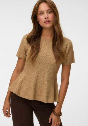 Mujer con cabello largo y castaño que lleva una camiseta de punto beige de manga corta y pantalones marrón oscuro, de pie frente a un fondo blanco liso.
