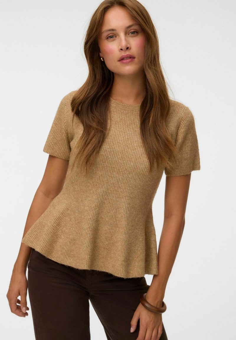 Femme aux longs cheveux bruns portant un haut en maille beige à manches courtes et un pantalon marron foncé, debout devant un fond blanc uni.