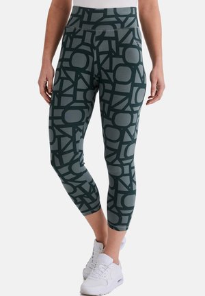 Frau trägt graue und schwarze, geometrisch gemusterte, gekürzte Leggings mit weißen Turnschuhen, steht vor einem einfarbigen Hintergrund.