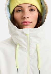 White waterproof jacket with a high collar, yellow drawstrings, and a blue lining. The model wears a light yellow ribbed knit beanie.